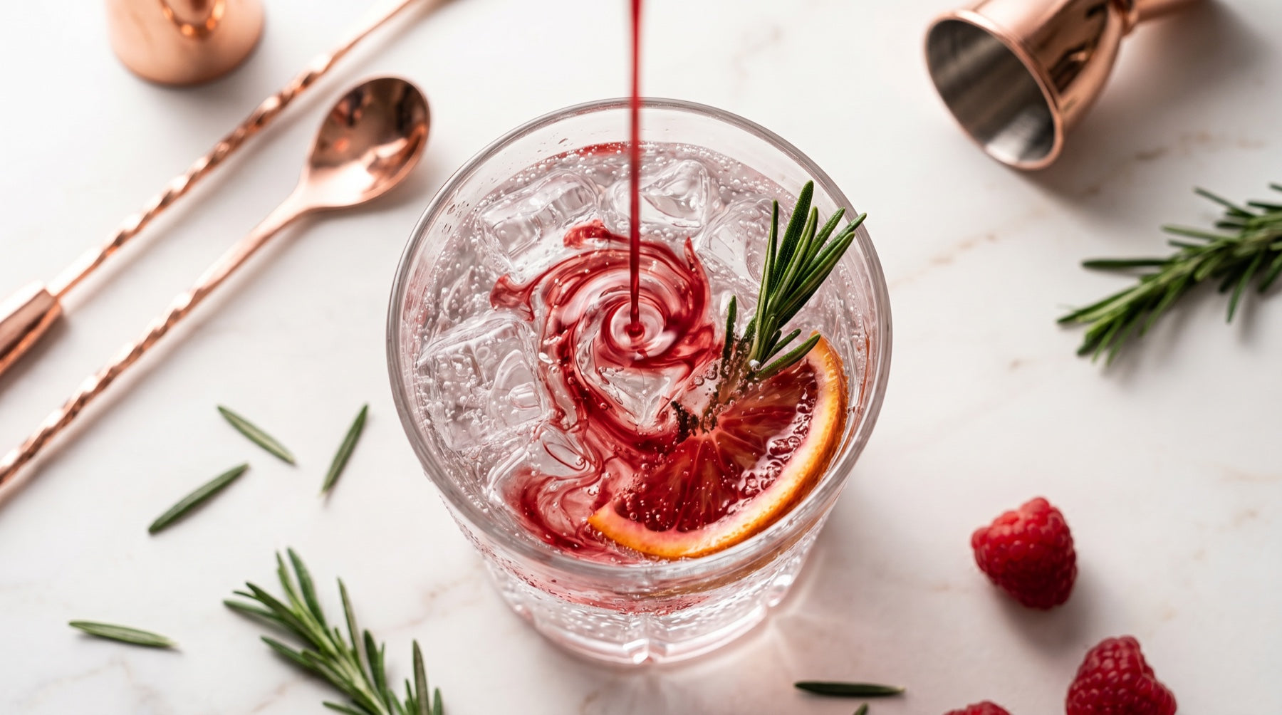 Overhead view of a crystal glass of sparkling water over ice with a blood-orange syrup pour, rosemary and fresh fruit on a marble bench